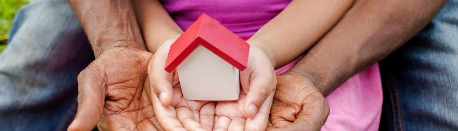 Handen van papa en pleegkind rond houten huisje: de pleegzorgtoeslag